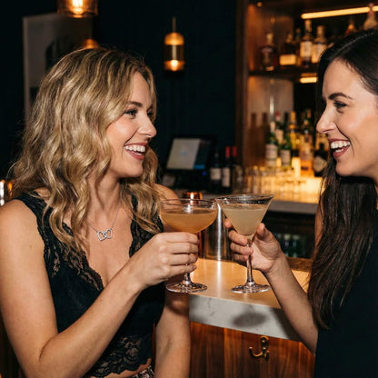 Best friends toasting with cocktails on a girls' night out, wearing the interlocking hearts friendship necklace.