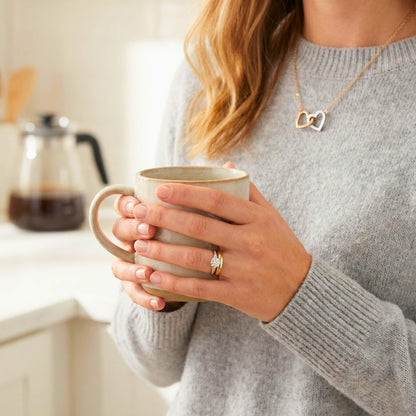 Close-up detail of wife wearing the interlocking hearts necklace while holding a morning coffee, showing everyday elegance.